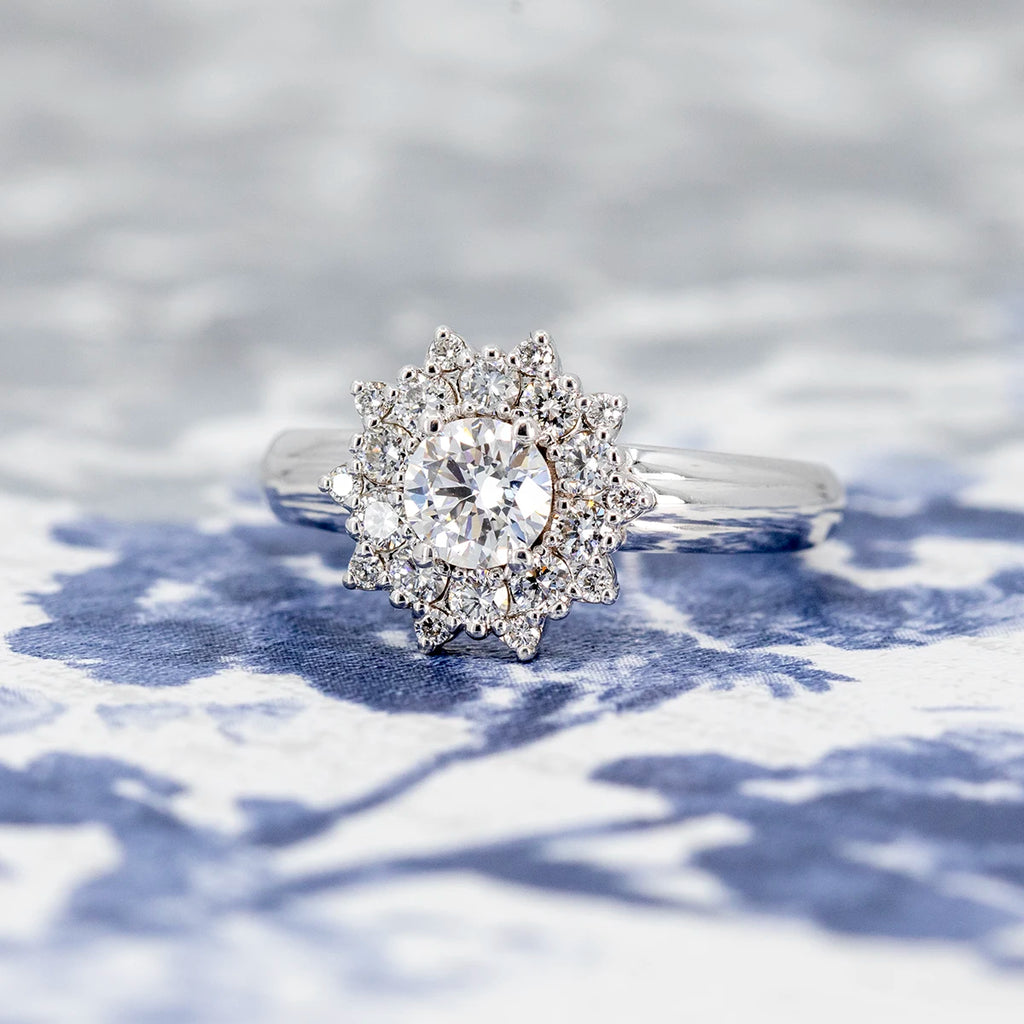 Starburst engagement ring in white gold with a central round brilliant diamond and a double halo of smaller diamonds, designed by Bena Jewelry. Photographed on a blue and white floral fabric background. Elegant handmade ring available at Ruby Mardi, Montreal.