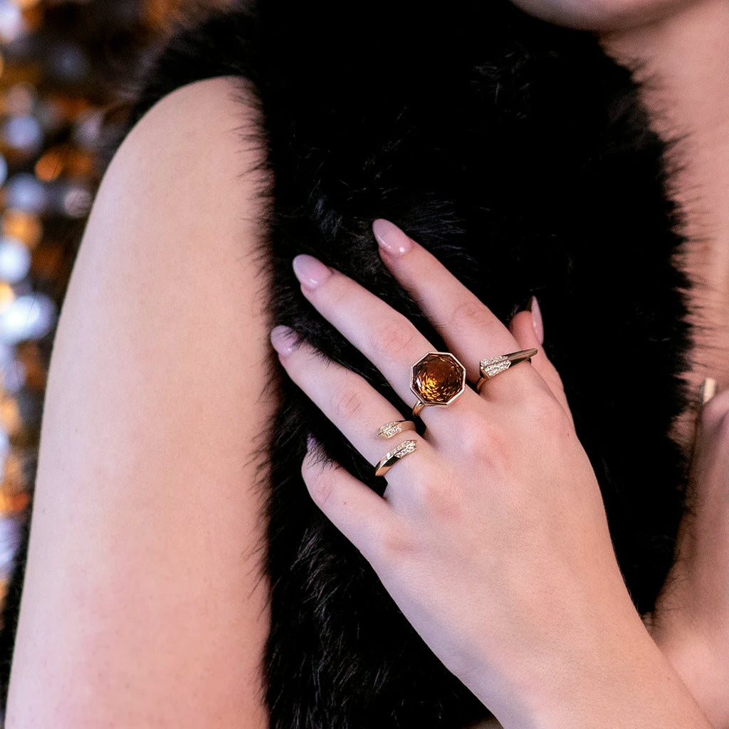 A close-up image showcasing a person's hand adorned with several gold rings, including one with a prominent amber-colored gemstone. The person is wearing a black fur-like garment, and the background features a blurred display of warm lights, emphasizing an elegant and luxurious style.