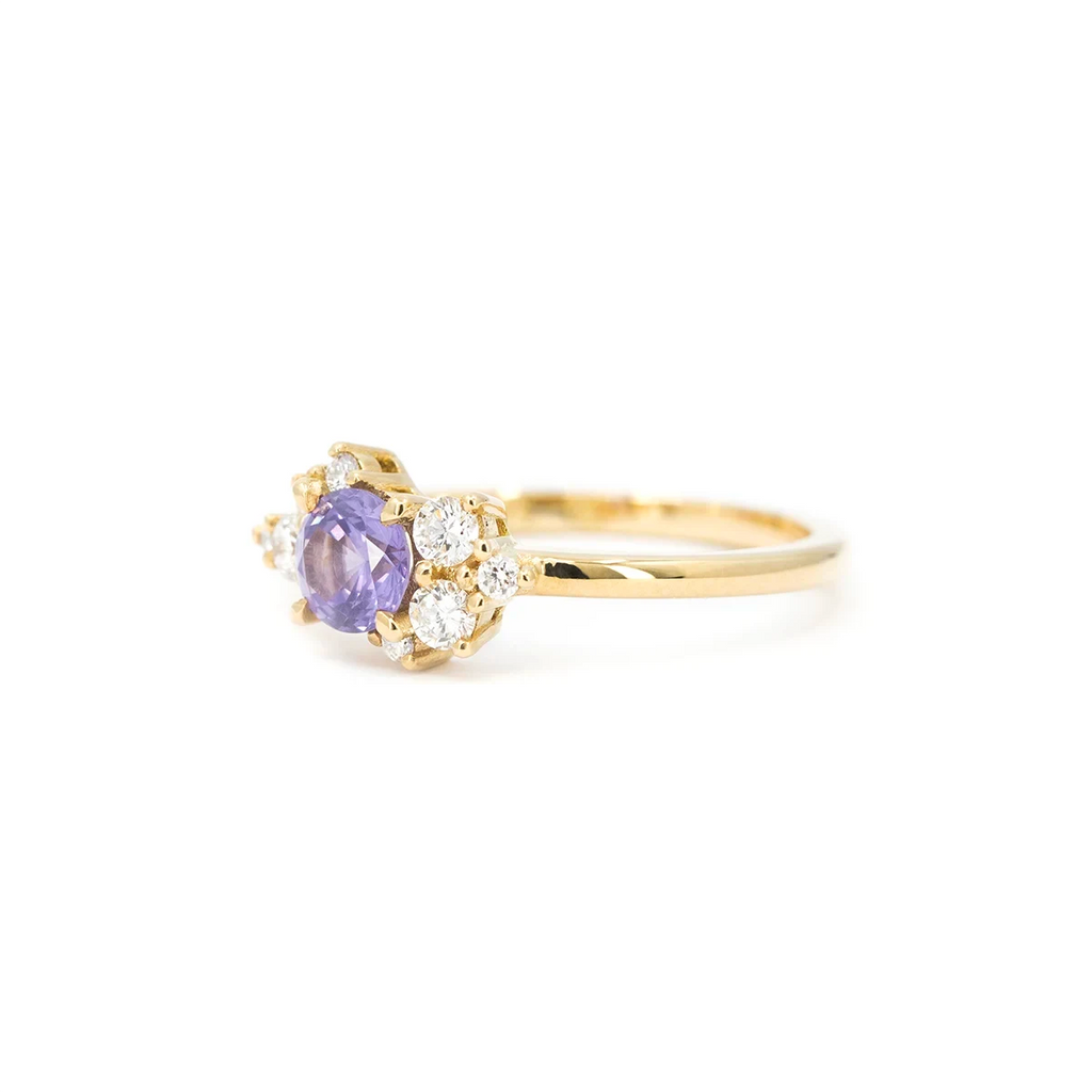 "A gold ring handcrafted by Justine Quintal, featuring a round purple sapphire center stone accented with asymmetrically arranged small round white diamonds, displayed on a white background.