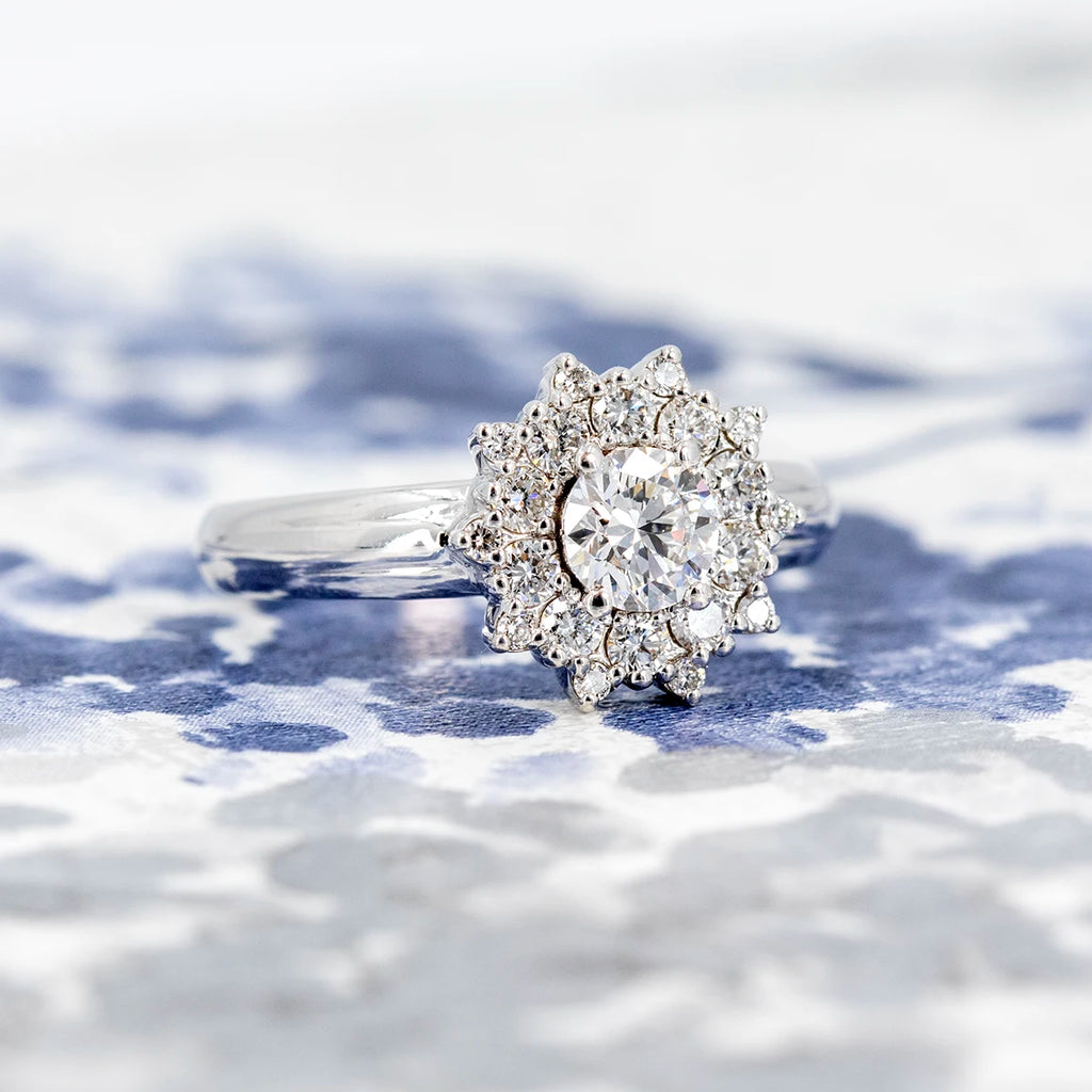 White gold engagement ring with round brilliant center diamond and intricate double diamond halo, designed by Bena Jewelry. Displayed on a blue and white floral fabric. Elegant fine jewelry available at Ruby Mardi, Montreal.