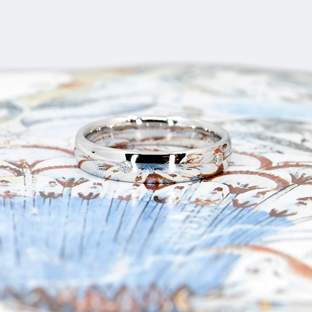 A white gold wedding ring with small diamonds on the band is seen photographed on an abstract blue and brown background that is reflected o the high polished ring. This original jewelry piece was handcrafted in Montreal by independent jewelry brand and store Ruby Mardi. We offer Canadian jewelry and designer jewels.