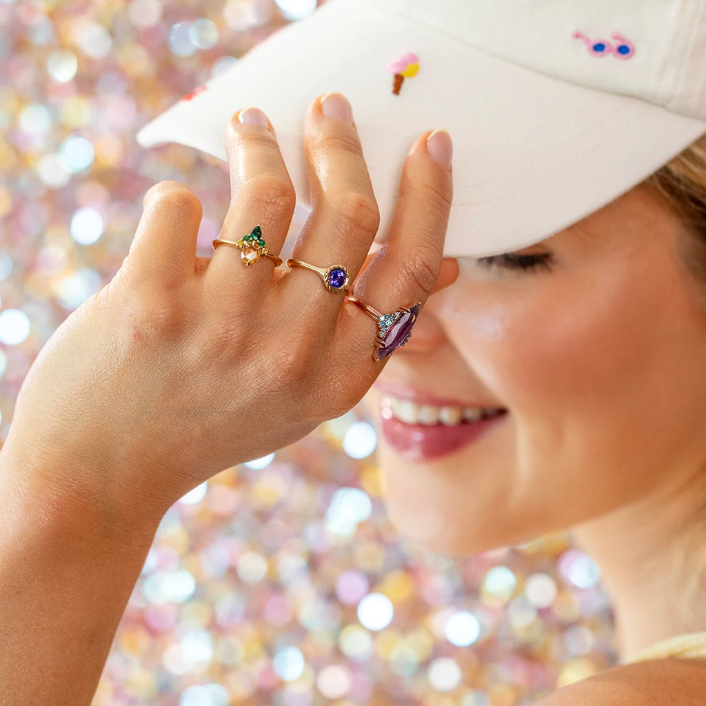 Smiling woman in a white embroidered cap showing her hand adorned with three vibrant gemstone rings in yellow gold: a pear-cut citrine with green accent stones, a round vivid blue sapphire, and a bold statement amethyst ring with geometric details. Playful and colorful fine jewelry styling captured in natural light.