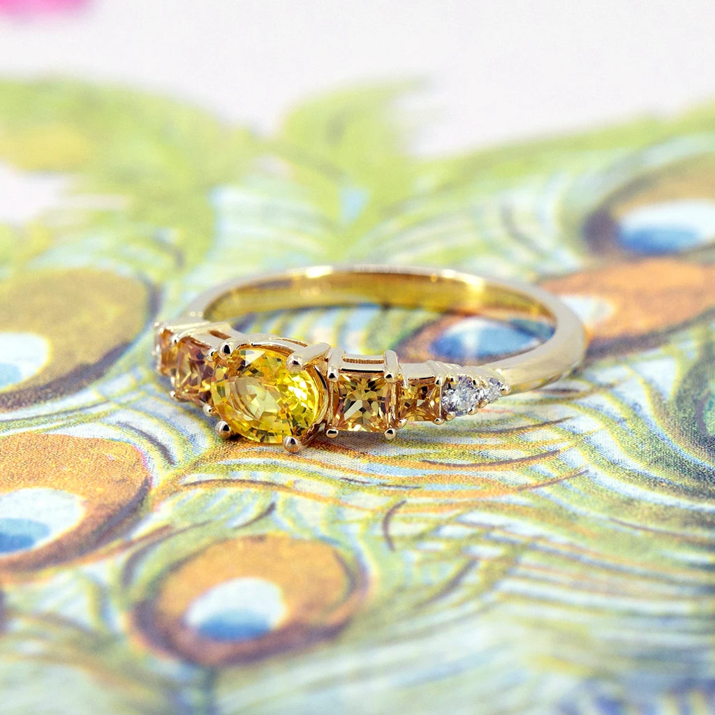 An engagement ring in yellow gold with a central oval yellow sapphires and 4 side princess cut orange sapphires, as well as diamond accents is seen photographed on an illustration of a peacock. This fancy bridal ring was designed and handcrafted in Montreal, Canada, by Ruby Mardi.