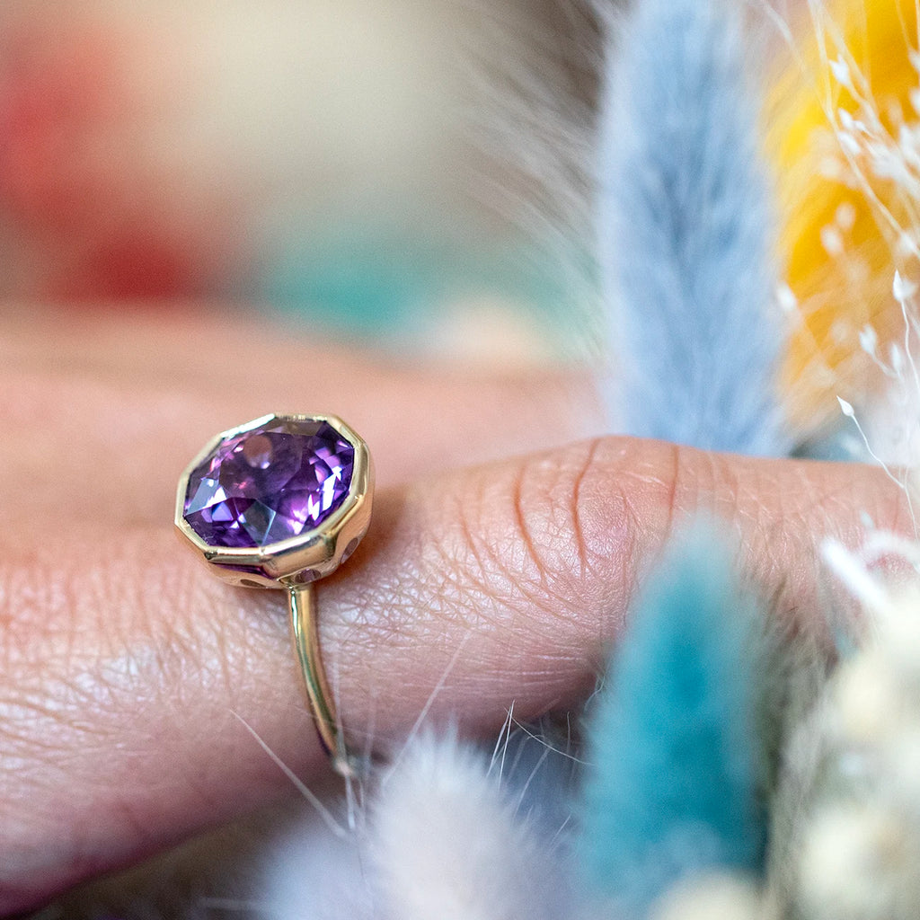 Finger wearing a unique montreal jewelry designer ring with a fancy shaped purple amethyst gemstone mounted in yellow gold in bezel setting on a multi color background