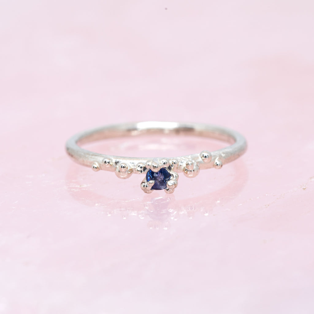Organic ring by Meg Lizabet made of sterling silver and featuring a small blue sapphire and silver granules. Seen photographed over pink quartz.