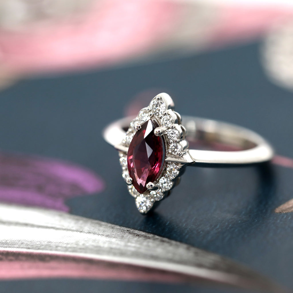 Engagement ring with a marquise shape raspberry-red garnet and a halo of brilliant diamonds, set in 14kt white gold. Seen here photographed on a dark romantic wallpaper. This luxury jewel was created and manufactured in Montreal by independent brand Ruby Mardi, which also represents Canada's top jewelry designers, as does Gem Breakfast in the USA.