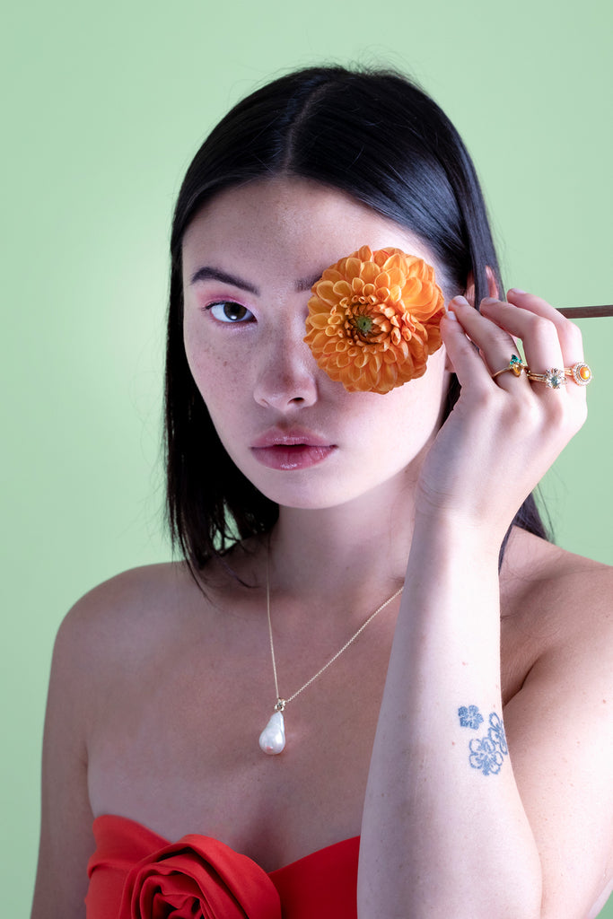 Beautiful photo studio of an asiatic girl hiding one of her eyes with an orange dahlia and wearing 3 designer gemstone rings (with sapphire, diamonds, opal, emerald and sapphire) and a baroque pearl necklace.