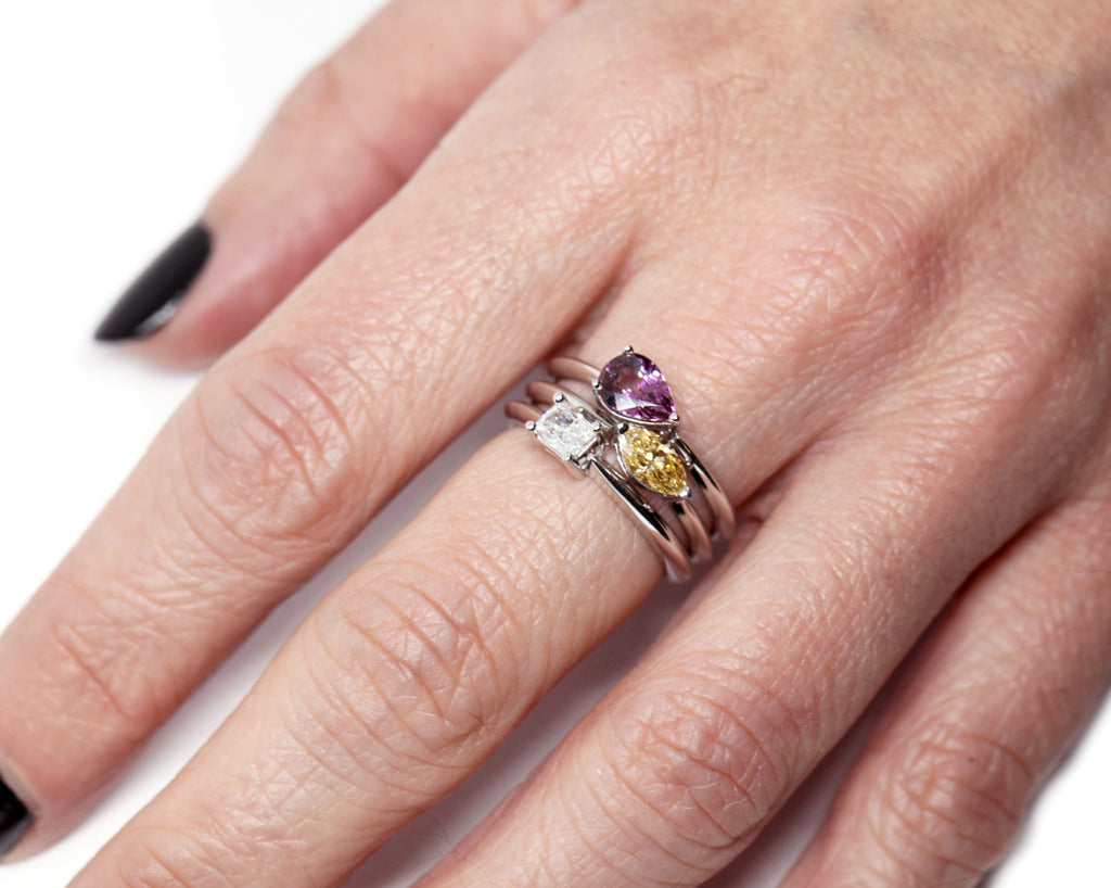 Close up on 3 white gold solitaire rings on a hand : one with a radiant diamond, one with a Malaya garnet, and the other one with a yellow sapphire. Our high end jewelry store offers custom jewellery designs services. 