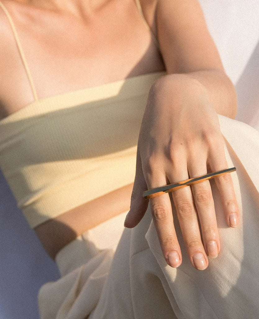 A statement double ring by Bena Jewelry, featuring an elegant geometric design. This modern piece is worn on two fingers, combining bold lines and a minimalistic style. The ring is photographed on a hand wearing light, casual attire, with a soft, sunlit background highlighting its striking appearance.