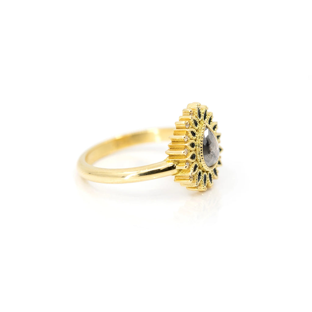 An 18-carat yellow gold ring is photographed in three-quarter view against a white background. An 18-carat yellow gold ring is photographed up close on a woman's finger with long pale pink nails. This is a unique designer ring featuring a pear-shaped salt-and-pepper diamond at its center, surrounded by several small black pear shapes in enamel. Completing the composition are 16 natural diamond accents. This alternative engagement ring is available only at Ruby Mardi.