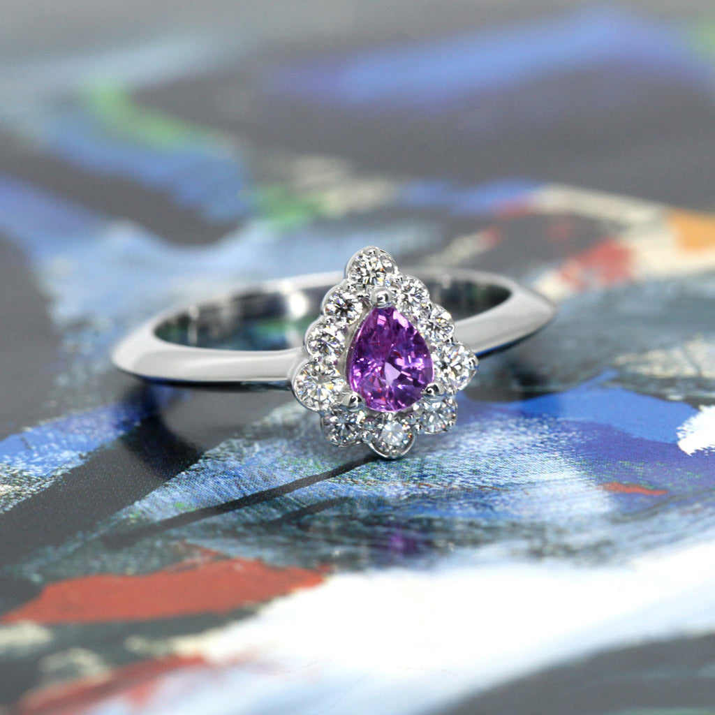 Pear Shape Purple Pinkish Sapphire Engagement ring with a diamond halo on a white background by Ruby Mardi, a fine jewelry gallery in Montreal Little Italy, close by Bijouterie Italienne, and Rosemont, Outremont, Villeray, Parc Extension, Mile End, Mile Ex districts. White gold gemstone ring, bridal jewelry, wedding ring, ethical gem. Bicolor Malaya Garnat. Ruby Mardi offers custom jewelry services in Montreal. Lab grown diamonds, natural diamonds, Canadian diamonds, ethical diamonds, ethical gemstones.