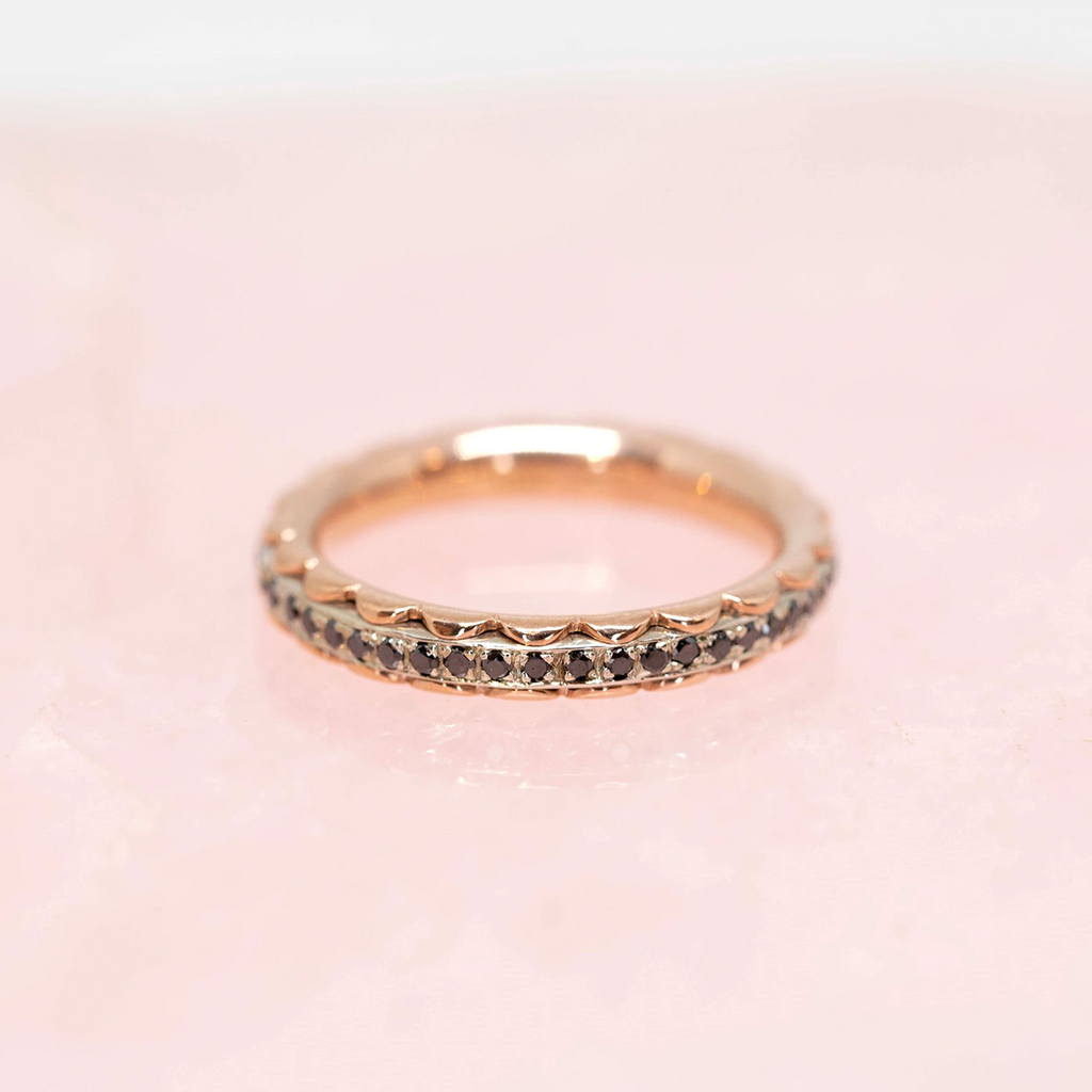 Handmade eternity wedding ring by fine jewelry designer Janine de Dorigny. Unique wedding jewelry made in rose gold with black diamonds. Set with small dark and refined precious gems, placed on a pink background. This ring is available at the Boutique Ruby Mardi jewelry store in Montreal's Little Italy.