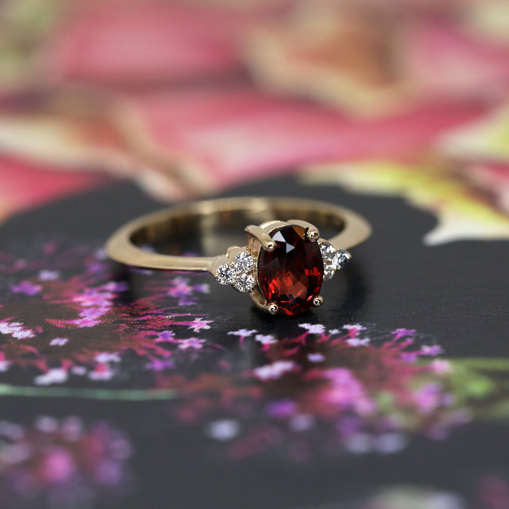 Close up on an engagement ring featuring a beautiful red garnet with six round brilliant diamonds and photographed on a flowery background. Find more fine jewelry and bridal jewels at boutique Ruby Mardi in Montreal.
