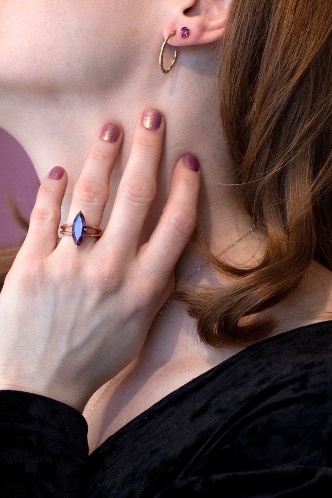 girl wearings a long amethyst marquise shape rose gold statement ring and two earrings with a sand cast yellow gold hoop and minimalist small oval shape red garnet studs on a purple background