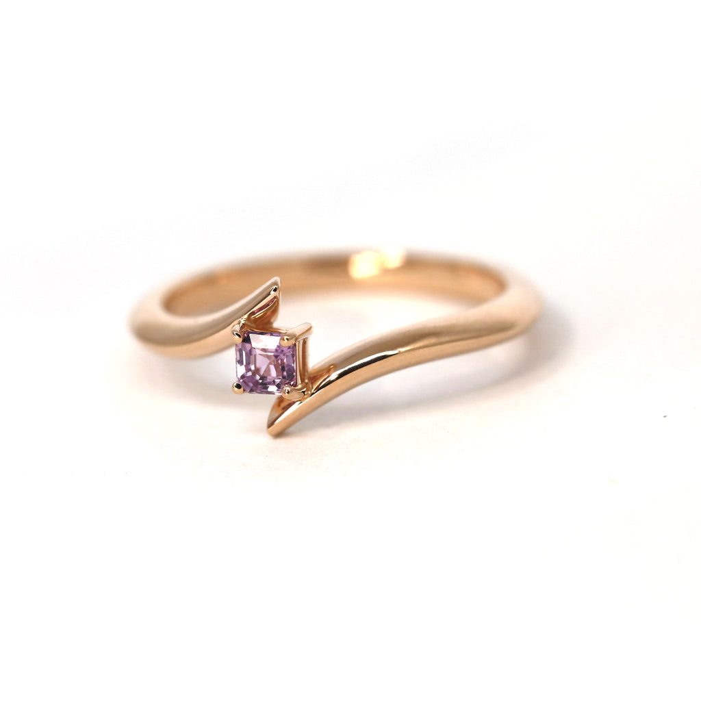 Twisted rose gold engagement ring with an emerald cut purple-pink sapphire photographed on a white background. More one of a kind jewelry available at Ruby Mardi, a jewellery store in Montreal’s Little Italy.