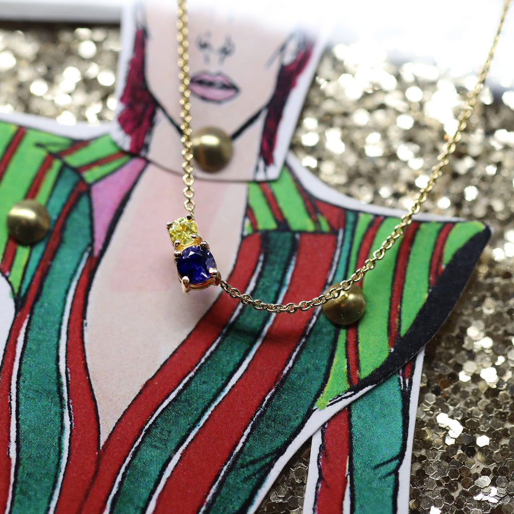Joli pendentif avec pierres précieuses jaune et bleue créé et fabriqué à la main par la créatrice de bijoux montréalaise Lico Jewelry et photographié au-dessus d’une poupée de papier de David Bowie. Disponible à la galerie de bijoux fins Ruby Mardi, dans la Petite Italie.