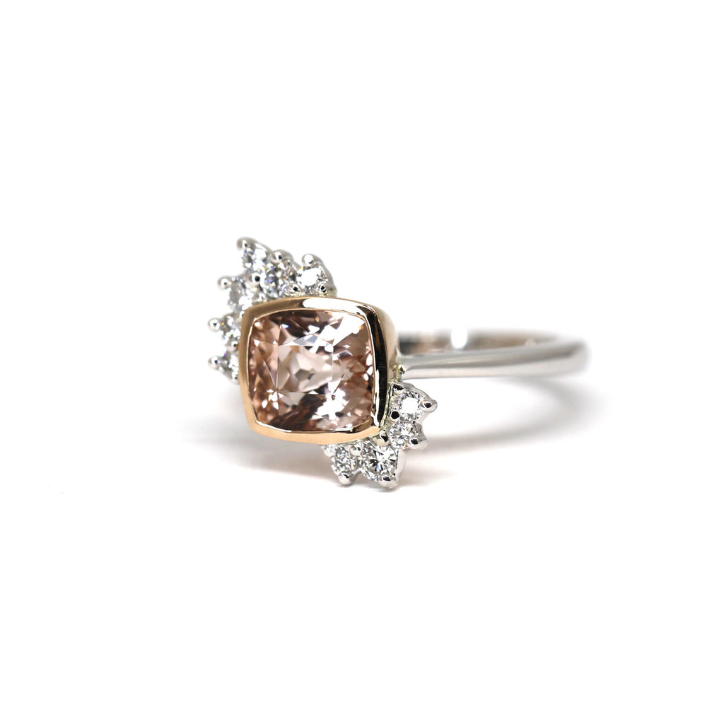 Side view of an engagement ring in white gold and rose gold with a cushion morganite and round brilliant diamond accents seen on a white background. It’s an asymmetrical ring with a feminine touch. This bridal jewel is only available at Ruby Mardi, in Montreal’s Little Italy or on the website. It is available to ship. 