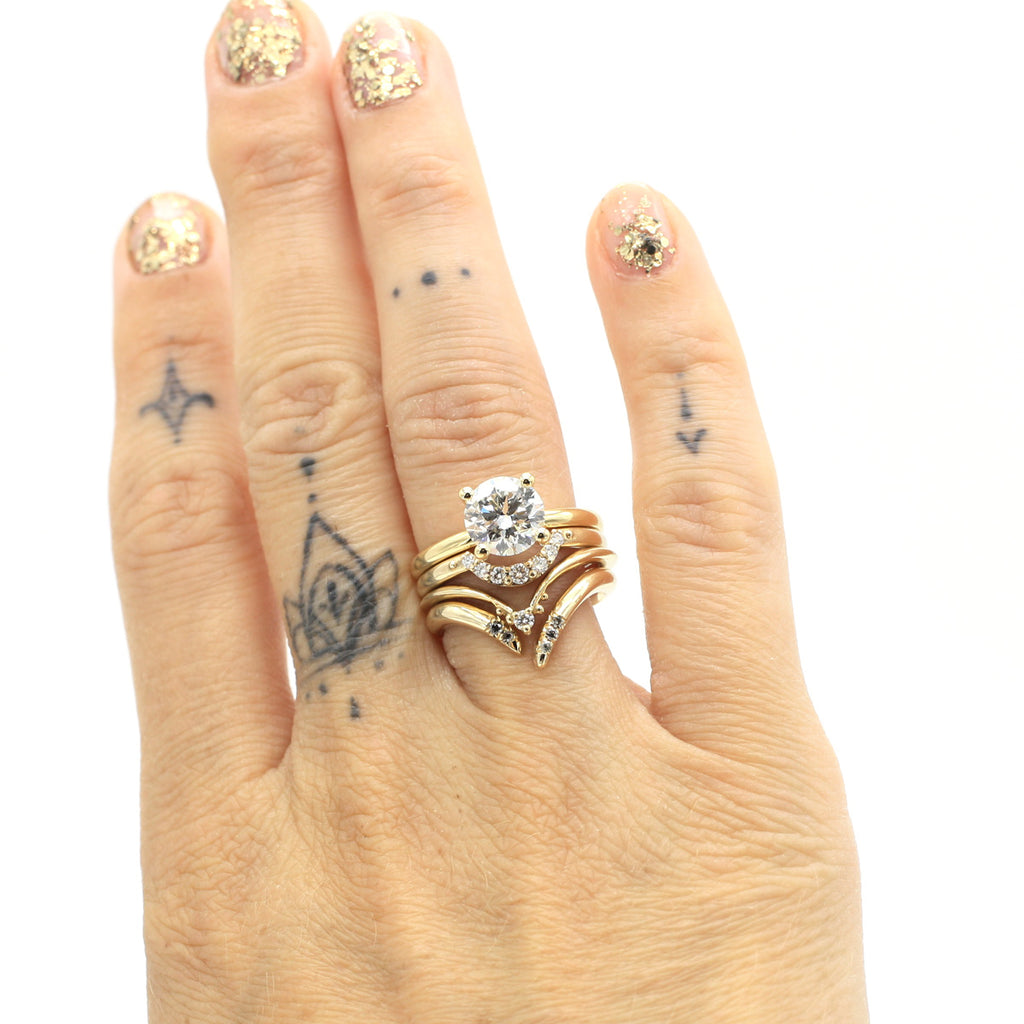 An ambitious stacking of precious ring seen on a hand with tattoos and glitters on the nails. Really precious rings in yellow gold with diamonds : One engagement ring with central round brilliant diamond, and 3 wedding bands matching with one another. 
