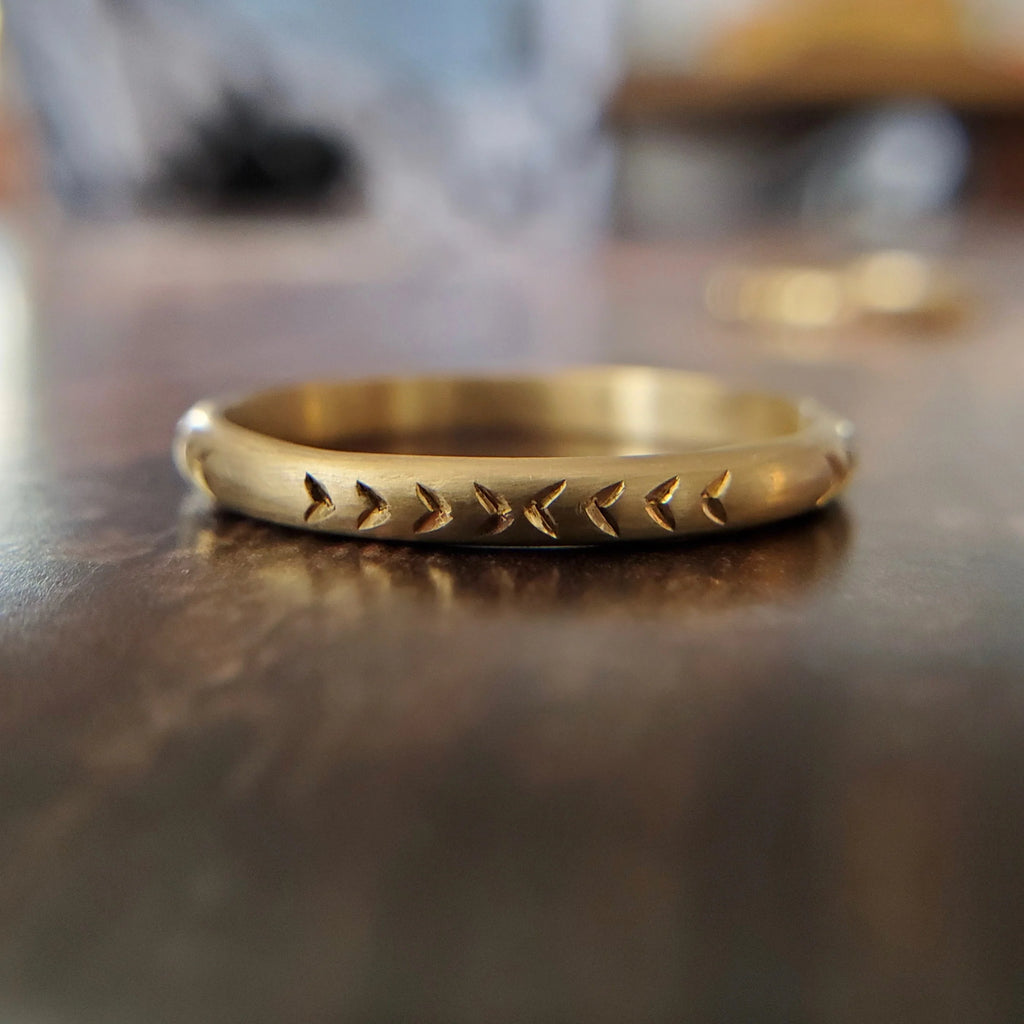 Gold band in 14k yellow gold with an hand engraved arrow pattern photographed on wood. Alternative wedding jewelry available at fine jewelry store Ruby Mardi, in Montreal’s Little Italy.