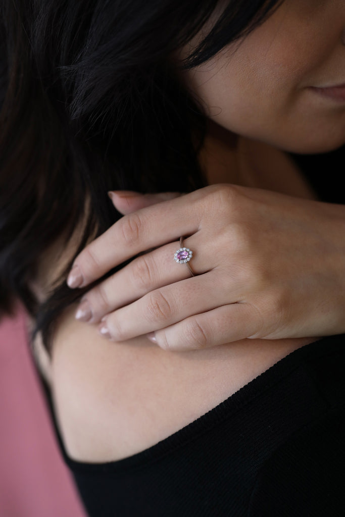 A lady wears an engagement ring with a romantic vibe and vintage feel. Oval pink sapphire and round brilliant diamond halo. Find it online or at our fine jewelry gallery in Montreal’s Little Italy.