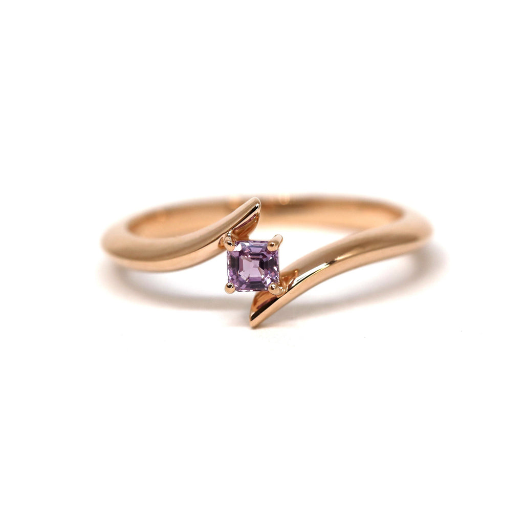 Twisted rose gold engagement ring with an emerald cut purple-pink sapphire photographed on a white background. A bridal piece of jewelry handmade in Montreal by local jewelry brand Ruby Mardi. 
