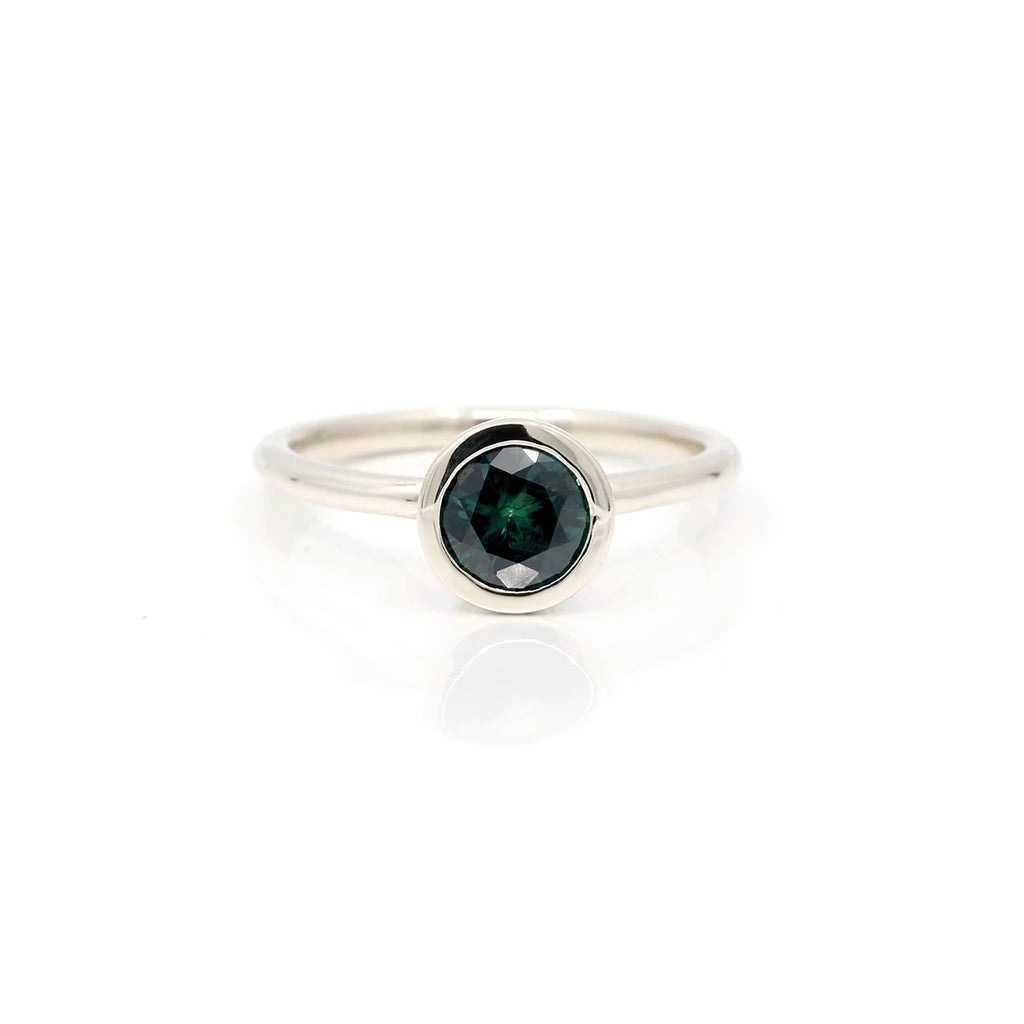 Perfectly round bezel set bluish-green sapphire bezel set in 18k recycled white gold ring is seen photographed on a white background. A piece of fine jewelry handmade in Toronto and available in Montreal at jewellery store Ruby Mardi.