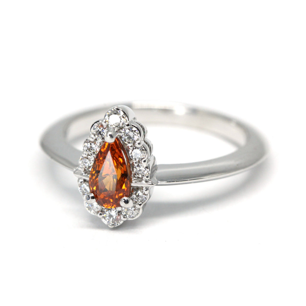 A spessartite garnet engagement ring with a diamond halo seen on a white background. This bridal piece of jewelry was handmade by Ruby Mardi, a fine jewelry gallery in Montreal Little Italy, close by Bijouterie Italienne. White gold gemstone ring, bridal jewelry, wedding ring, ethical gem. Ruby Mardi offers custom jewelry services in Montreal.