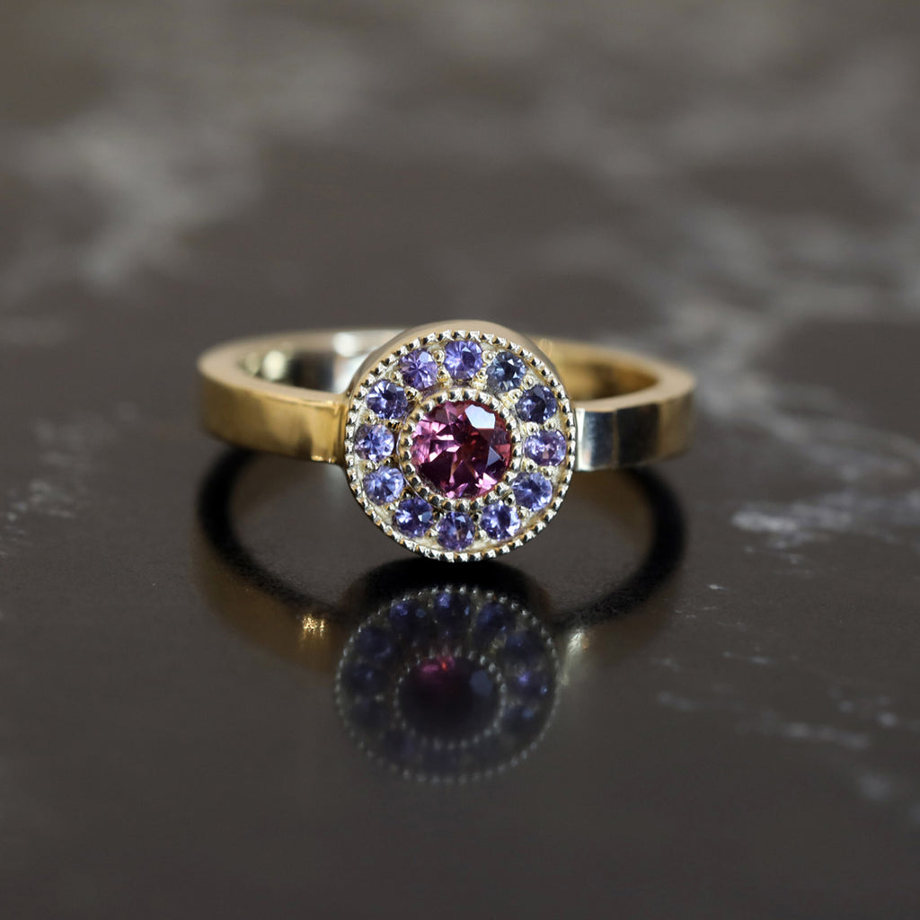 Front view on a bright light of a gold gemstone ring with raspberry spinel and a blue-violet sapphires halo. Beautiful jewelry handmade by Cecilia Lico and available at Ruby Mardi, a high end jewelry store showing the work of talented Canadian designers. 