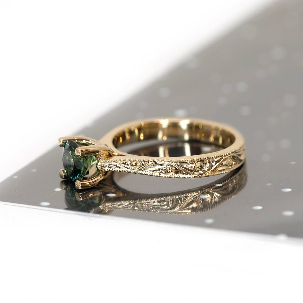 Side view of an engagement ring photographed on a black and white background. The yellow gold bridal ring features a round teal sapphire, raised in a classic fashion to easily accommodate a wedding band. The ring features hand-engraved scrolls. This bridal jewel was handcrafted by Deborah Lavery. Ruby Mardi represents this jewelry artist, as well as several other talented independent jewellery designers, as Gem Breakfast does in the USA.