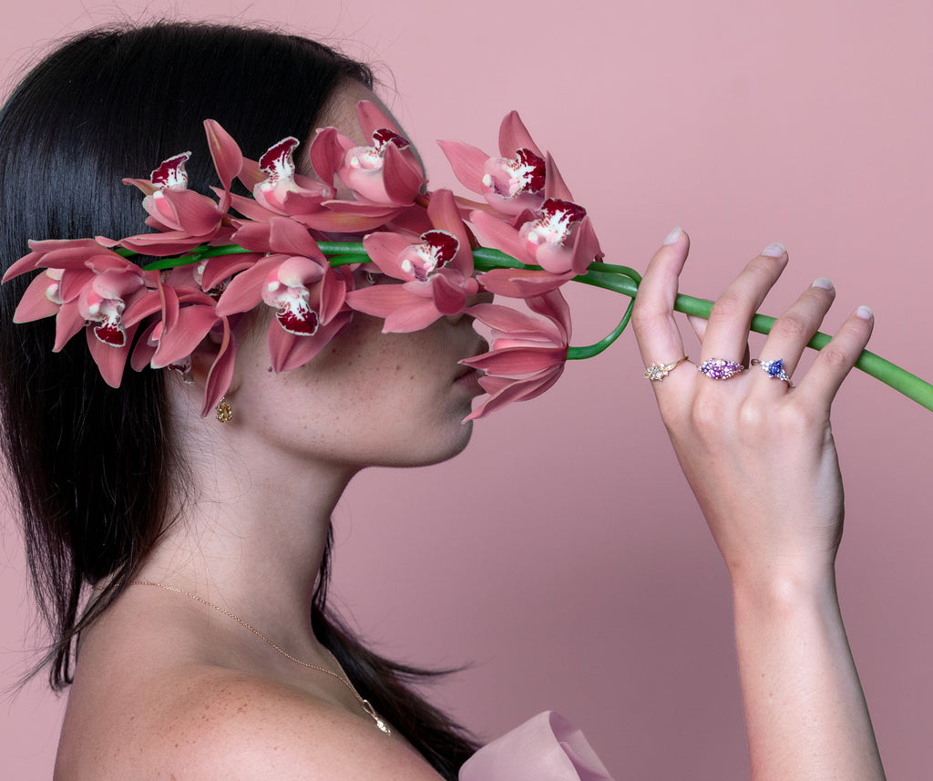 Artistic studio shot of a dark-haired Asian girl hiding her face with a large stem of purple-pink orchids. She holds the stem with one hand, wearing three designer engagement rings: an all-diamond ring, a ring with pink sapphires and diamonds, and a white gold ring with a central tanzanite. All these wedding rings are offered by independent Canadian jewelry store Ruby Mardi.