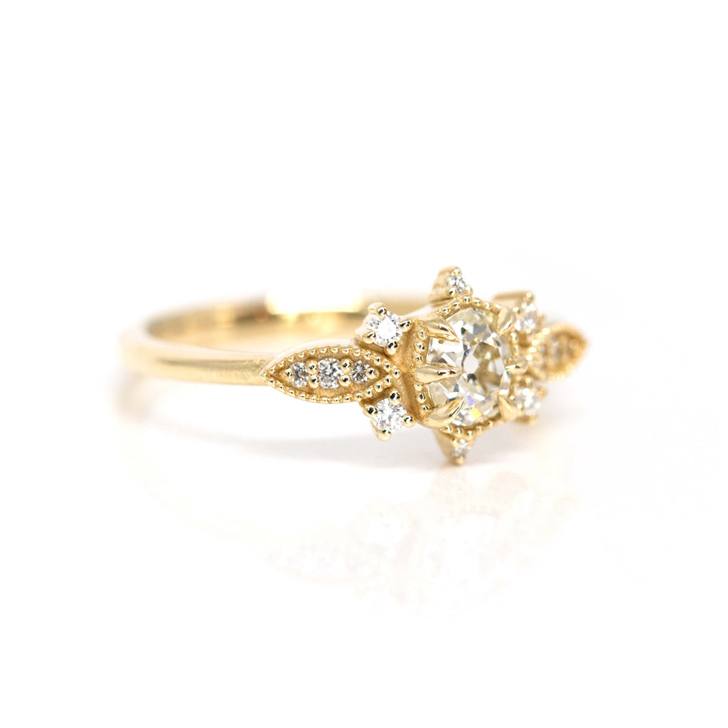 A beautiful engagement ring in solid  yellow gold with 13 natural diamonds is seen photographed on a white background. A work of art handmade by Nadia Werchola in Toronto, and exclusively available at Ruby Mardi in Montreal. The central diamond is old-mine cut, and there are many multigrain details that give a vintage vibe to the bridal jewel. 