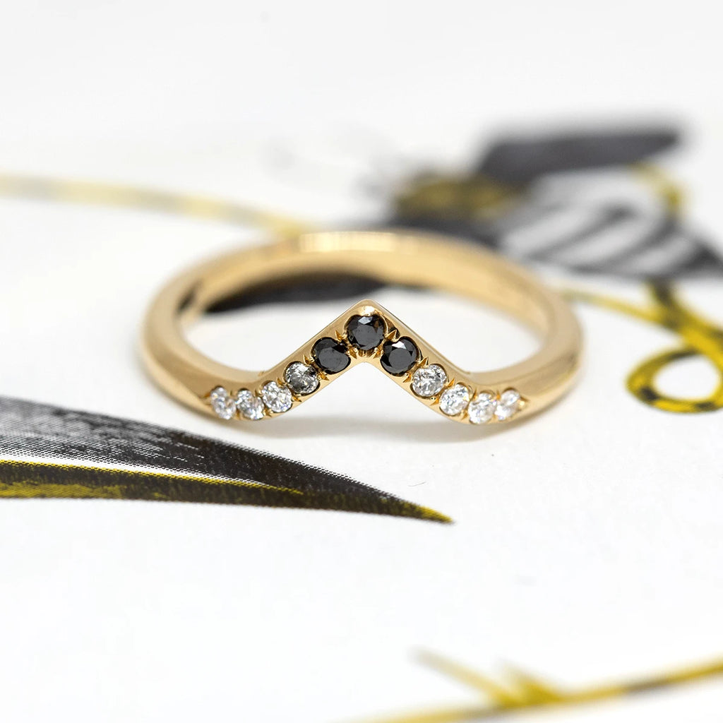A curved band in yellow gold featuring eleven diamonds in a gradation from white to black, photographed against an abstract yellow and black background. A wedding band handcrafted by Yuliya Chorna available at Ruby Mardi, a fine jewelry store showcasing the work of talented Canadian jewelry designers.
