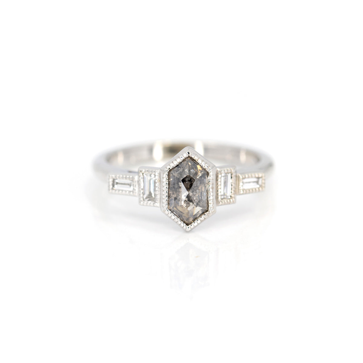 An art deco-style white gold ring is photographed against a white background. This right-hand jewel or engagement ring features a central hexagonal salt-and-pepper diamond, bezel set with miligrain detail. On the sides, 4 natural baguette diamonds, also bezel set with miligrain detail, give the set a retro charm. This unique ring was designed by Emily Gill in Toronto, and is available at the Ruby Mardi luxury jewelry store.