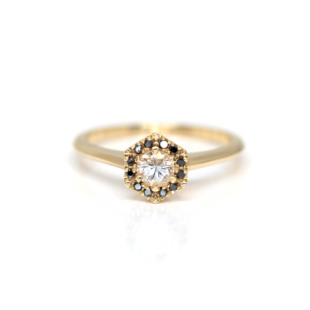 A designer engagement ring is seen photographed on a white background: a central round white sapphire is surrounded by a halo of black diamonds nested in an hexagonal shape. The ring is in yellow gold, it is an original design from Liane Vaz.