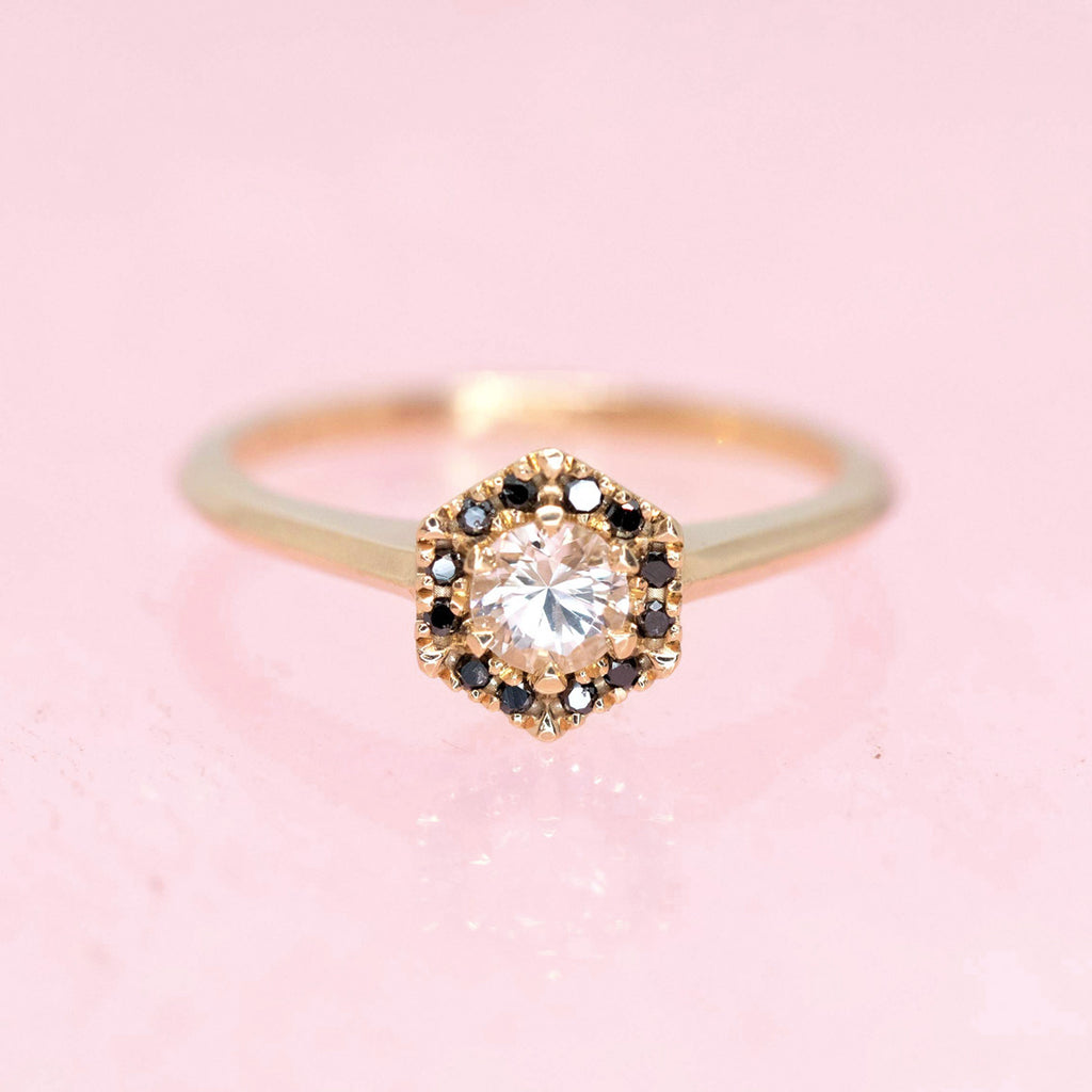 custom made engagement ring montreal with round white sapphire black diamond halo yellow gold ring on a pink background