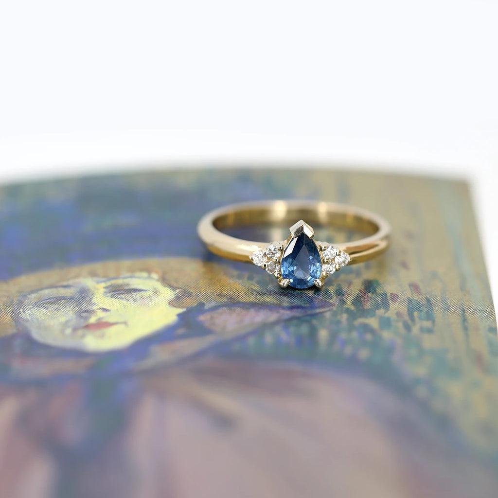 Gold ring with a pear blue sapphire and diamond accents, photographed on an artistic background. This one-of-a-kind engagement ring is available in Montreal and online, at jewelry store Ruby Mardi.