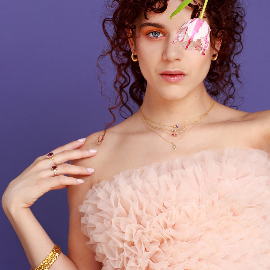Young woman with curly hair wearing several pieces of fine jewellery: bangle bracelets, gold rings with amethysts, 18-carat yellow gold hoop earrings and various necklaces with precious stones.