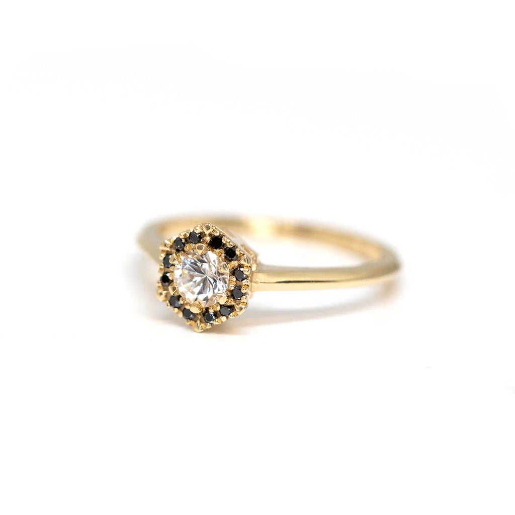 Handcrafted designer ring photographed against a white background, viewed from three-quarters up. The ring features a halo of black diamonds in a hexagonal shape, and a central round colorless sapphire. The ring is in yellow gold. It was handcrafted by designer Liane Vaz and is available at the best jewelry store in Montreal and Canada: Ruby Mardi.