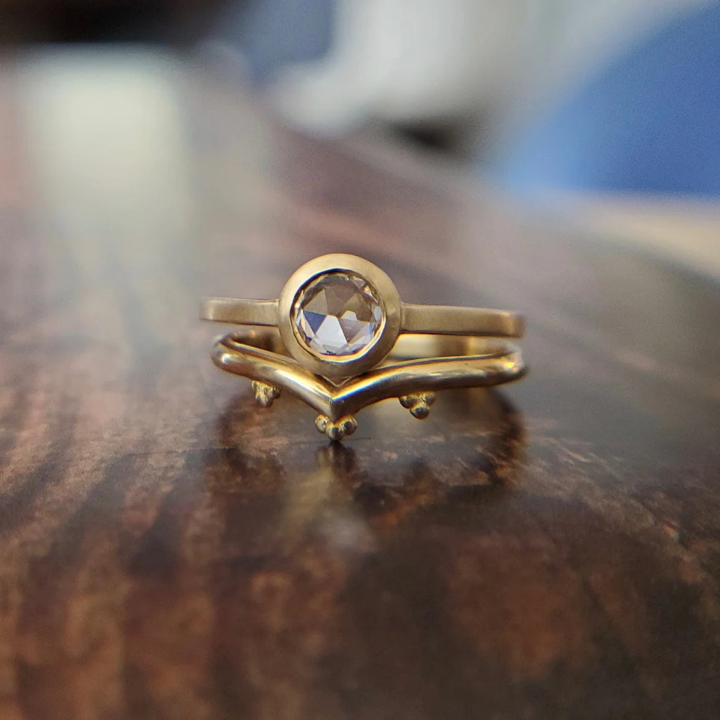 Two brushed gold rings seen stacked and photographed over a wood table. One ring features a round rose cut white sapphire, and the other is a v shape wedding band with chevrons. Both jewellery pieces were handmade in Toronto by designer Arsaeus Designs and are available at fine jewellery store Ruby Mardi, which sells the best engagement rings in Montreal.