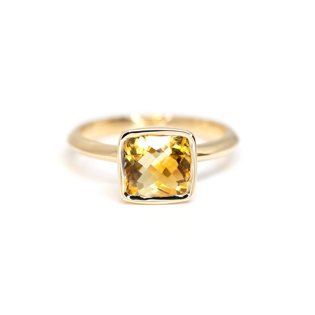 A shiny yellow gold ring with a bezel setting featuring a cushion cut citrine, seen on a white background. This ring was handmade and designed by Bena Jewelry in Montreal.