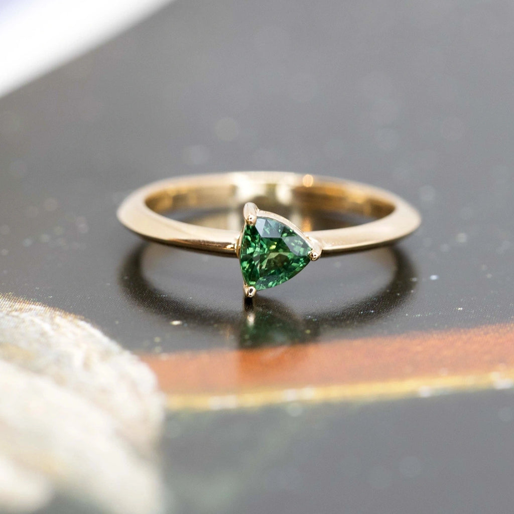 Engagement ring with a trillion-shaped green sapphire, this bridal ring is fine jewelry made in Montreal by Ruby Mardi. Yellow gold jewel with a deep green natural gemstone is placed on a dark background.