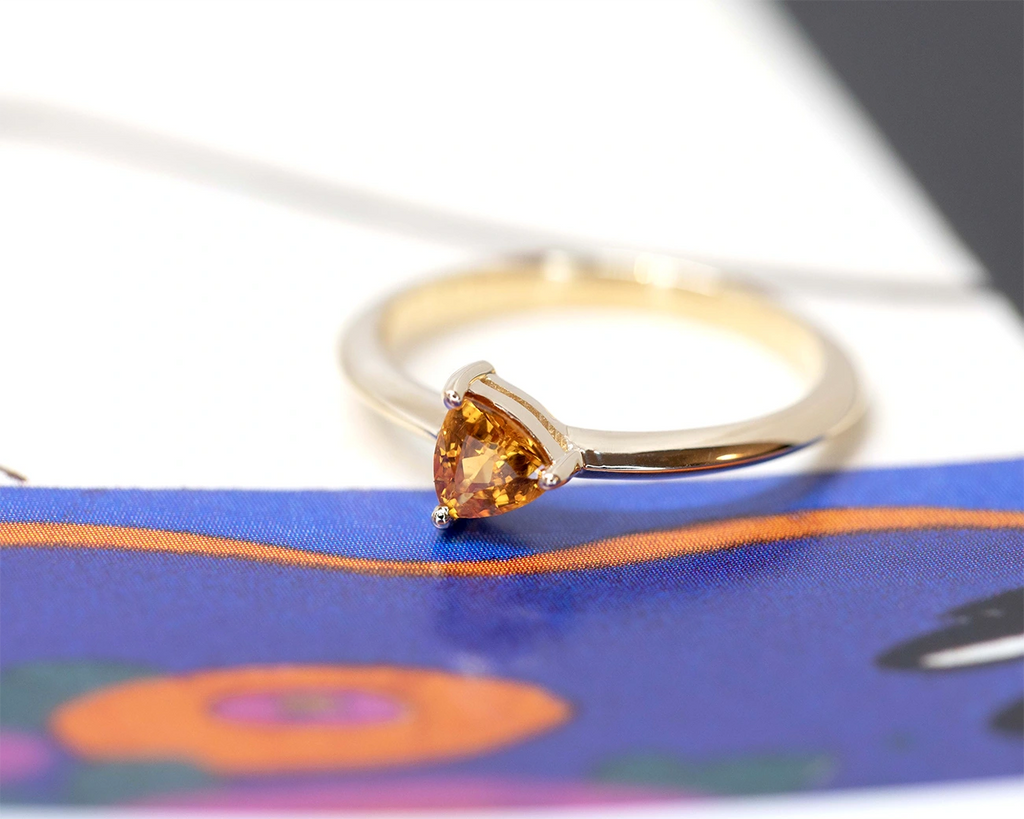 Side view of a trillion shape orange sapphire ring. A minimalist design for this custom made piece of jewelry handmade in Montreal by designer Bena Jewelry that is seen here on a funky background.
