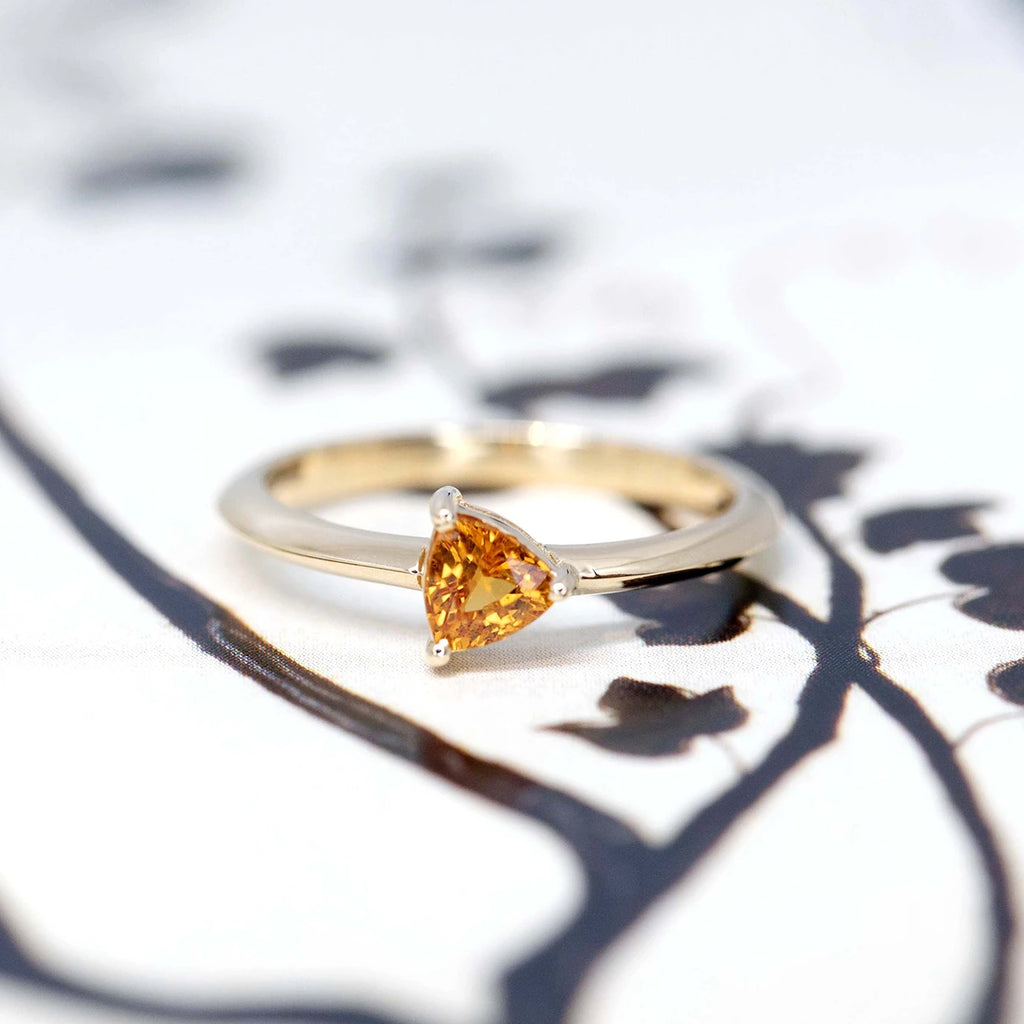 Front view of a custom made minimalist bridal ring featuring an orange trillion shape sapphire handmade in Montreal by designer Bena Jewelry, seen on a black and white background.