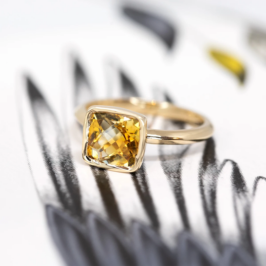 Side view of a yellow gold ring with a bezel setting featuring a cushion cut citrine, seen on an abstract black and white background.